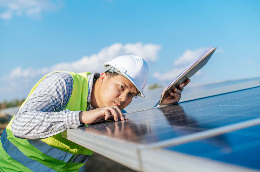 Young Asian Inspector Engineer man working at solar farm, Technician or supervisor male in white helmet Checking operation of sun and photovoltaic solar panel in station